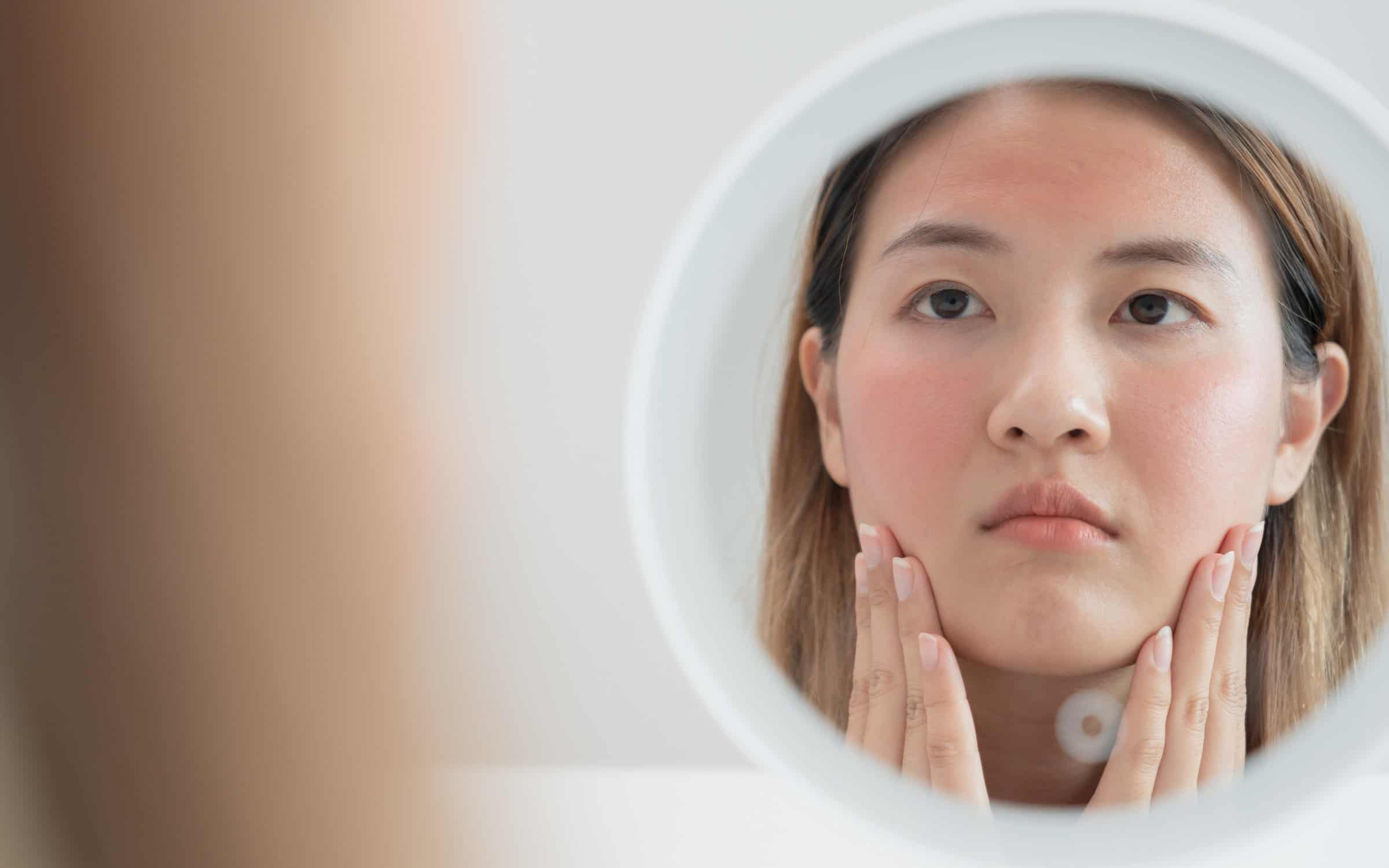 The image depicts a young Asian woman closely examining her face in a circular mirror, cupping her jawline and inspecting her skin.