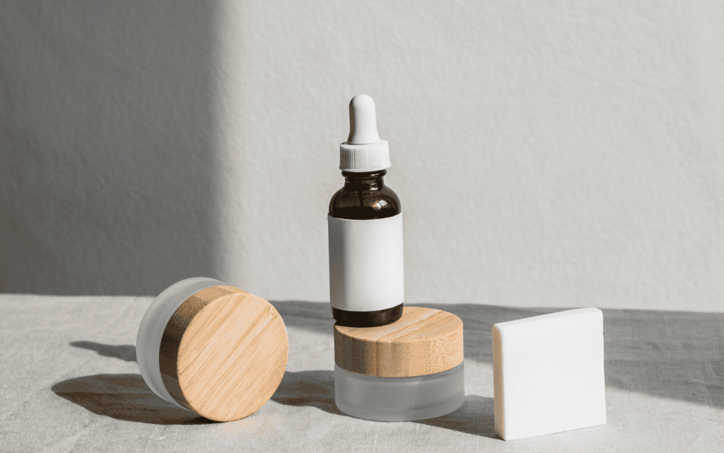 Glass dropper bottle and two cosmetic jars with wooden lids next to a blank rectangular box, all placed on a light fabric surface against a neutral background.