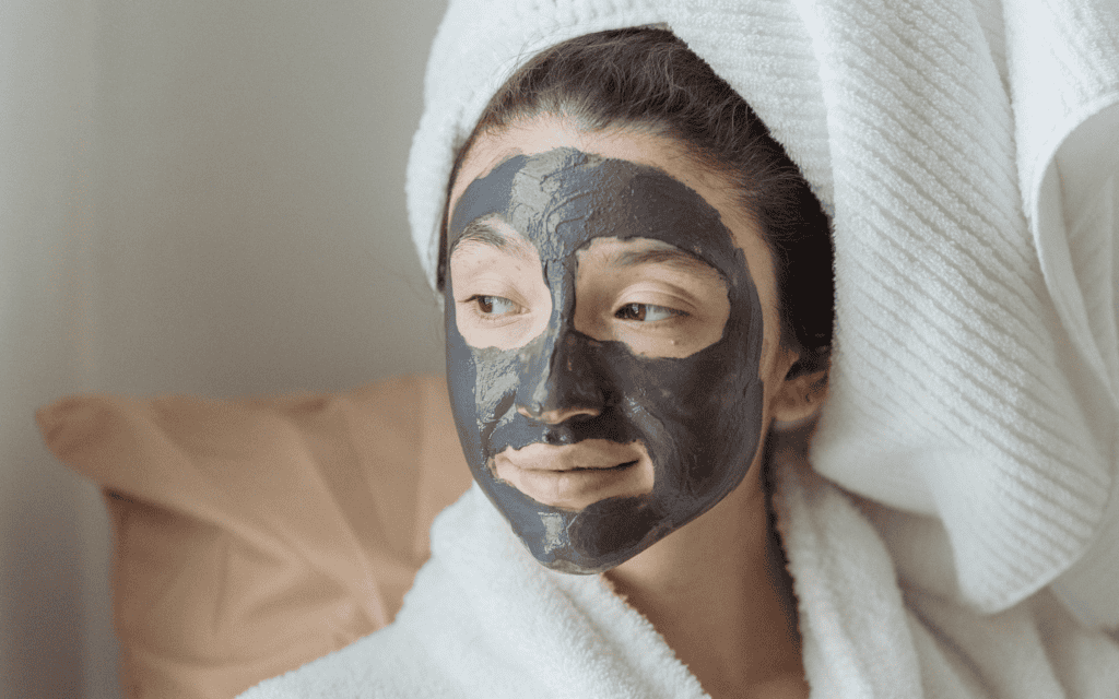 A person wearing a white bathrobe and towel on their head has a black facial mask applied, looking to the side while sitting on a bed with a brown pillow.