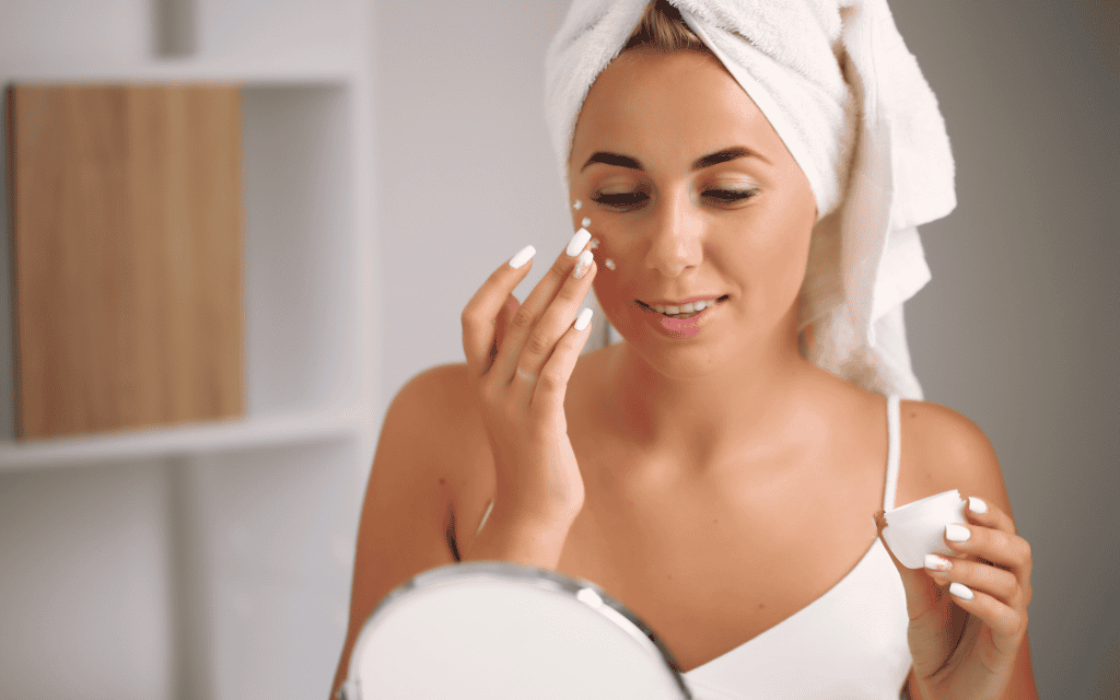 A woman with a towel wrapped around her head applies cream to her face while looking into a mirror.