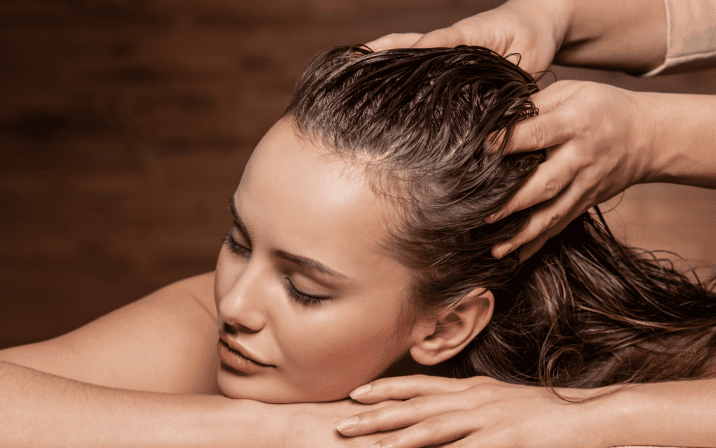 A person with wet hair receives a head massage while resting their arms and head on a surface, eyes closed in relaxation.