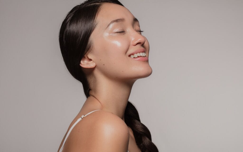 A woman with long dark hair in a braid smiles with her eyes closed, showing clear, glowing skin against a plain light background.