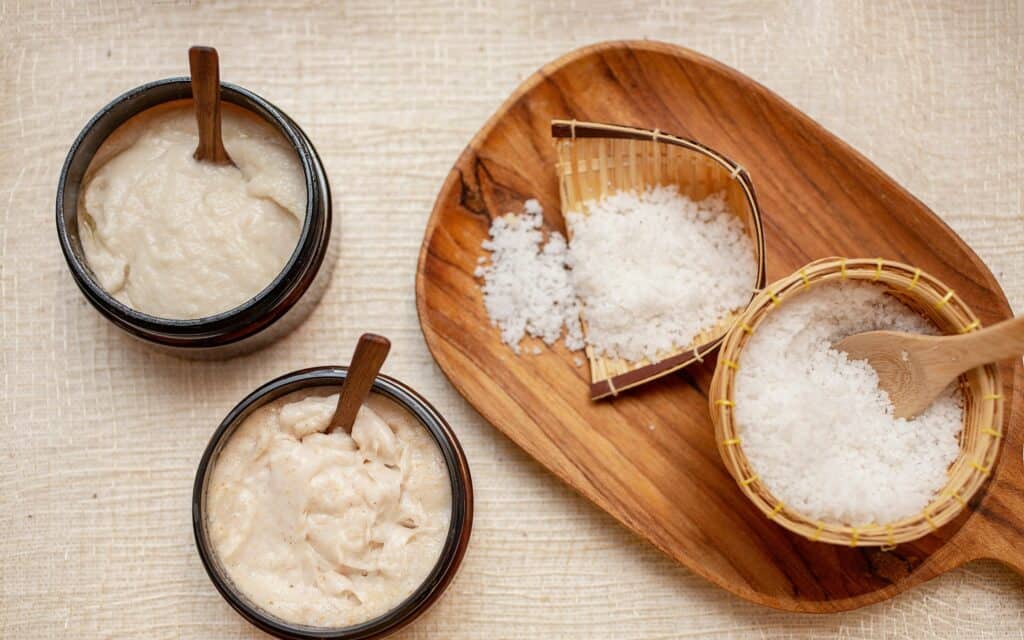 Two bowls with creamy mixtures each have small wooden spoons; a wooden tray holds a round bowl and two baskets filled with coarse white salt and a wooden spoon.