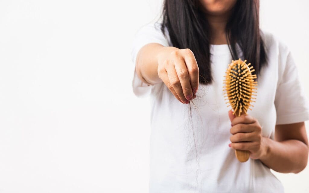 A woman holding a hairbrush in one hand and strands of fallen hair in the other, with her face partially out of frame.