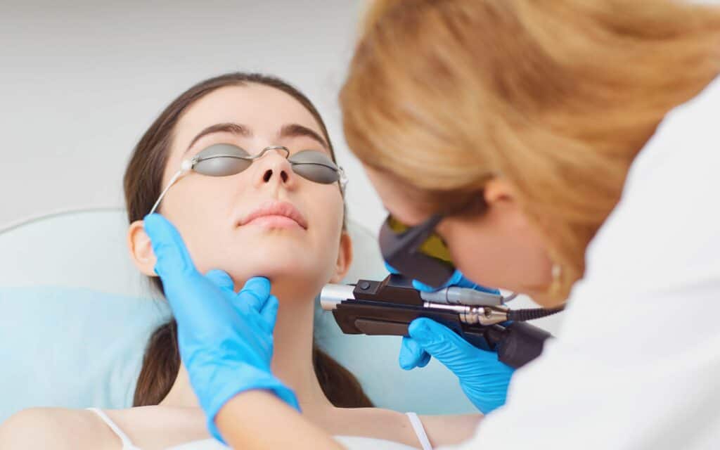 A woman wearing protective goggles receives a skin treatment with a handheld laser device from a gloved professional.