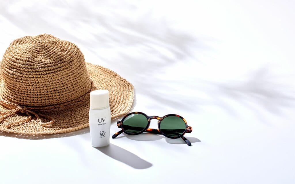 A straw hat, UV sunscreen bottle, and tortoiseshell sunglasses are arranged on a white surface with soft shadows.