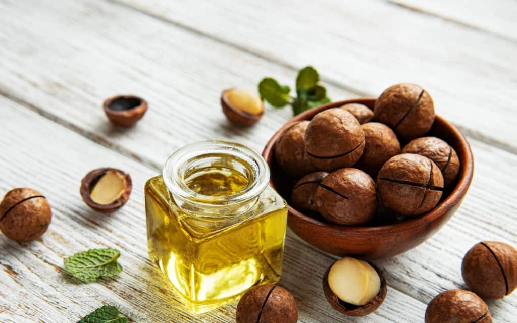 Junto a un cuenco de madera lleno de nueces de macadamia, algunas sin cáscara, sobre una superficie de madera blanca con ramitas de menta, hay un pequeño tarro de cristal con aceite de macadamia.