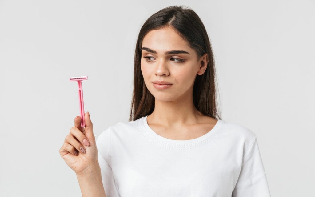 A woman in a white shirt holds a pink razor and looks at it, standing against a plain light background.