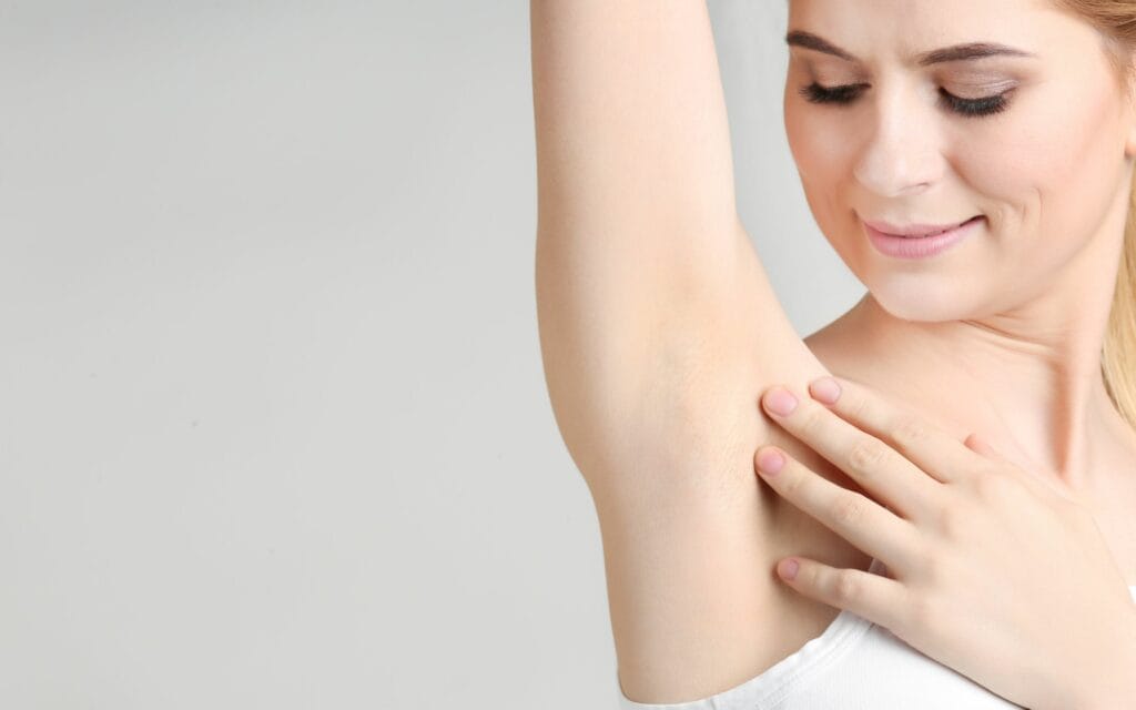 A woman in a white tank top examining or touching the skin of her armpit while looking down and smiling slightly.