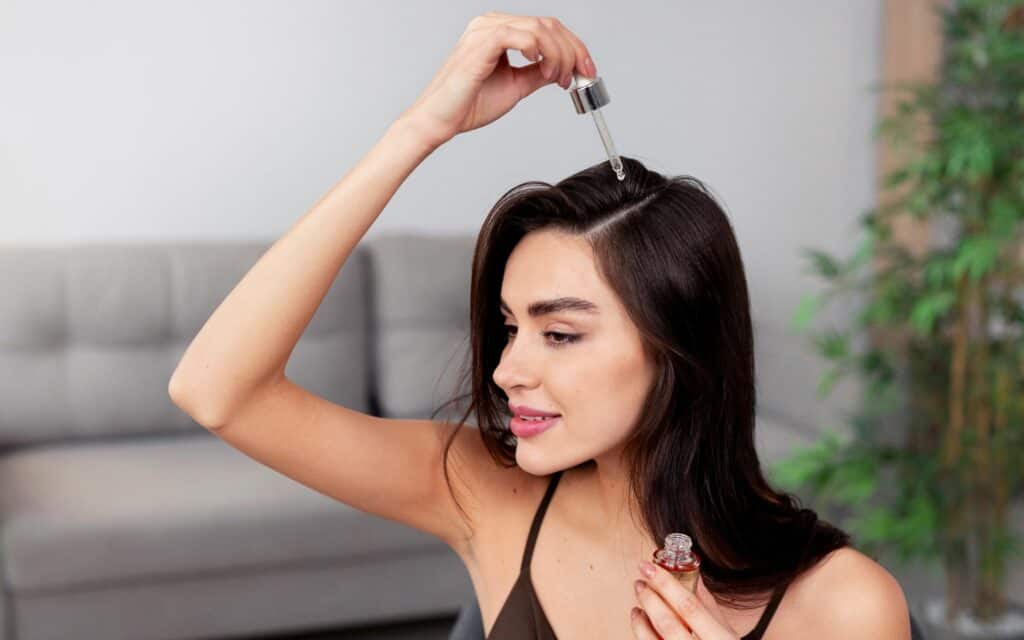 A woman applies hair serum to her scalp using a dropper, sitting indoors with a blurred sofa and plant in the background.