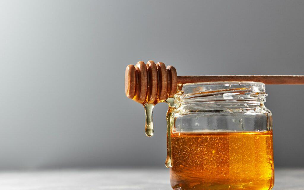 A wooden honey dipper rests on a glass jar filled with honey, with honey dripping from the dipper against a plain gray background.