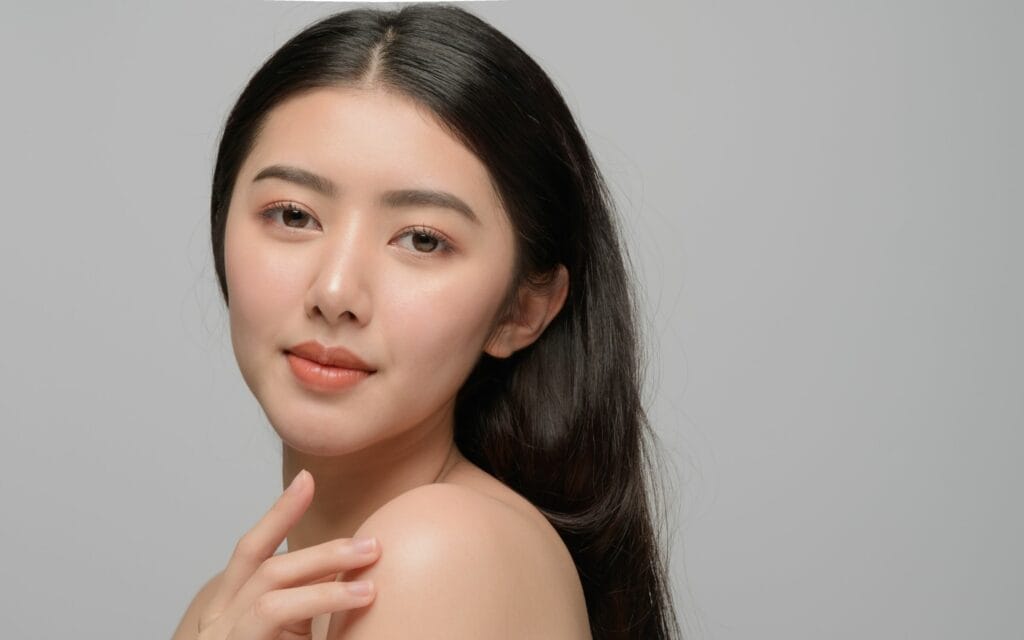 A young woman with long dark hair poses against a plain light gray background, looking at the camera with a neutral expression and her hand touching her shoulder.