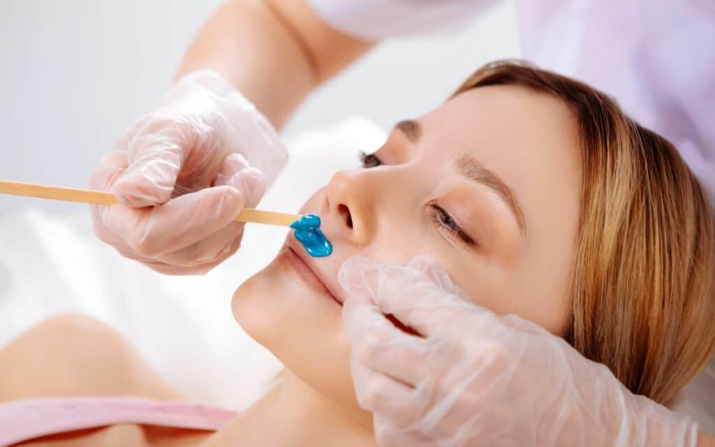A person wearing gloves applies blue wax to a woman's upper lip with a wooden spatula, preparing for a hair removal treatment.