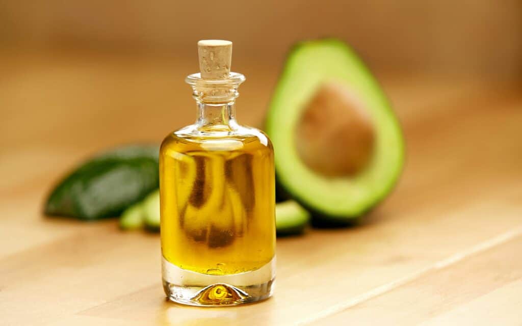 A glass bottle of avocado oil with a cork sits on a wooden surface, with a halved avocado in the background.